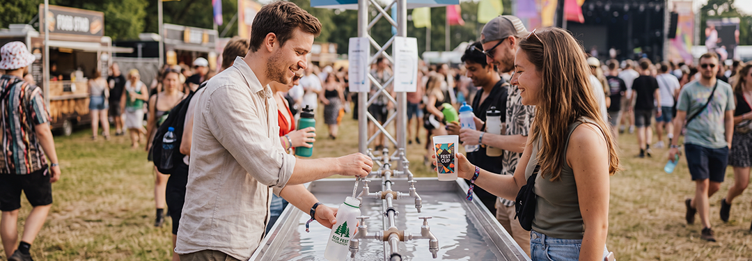 A man and woman at an outdoor drinks station, with the woman holding a printed cup beside several taps.